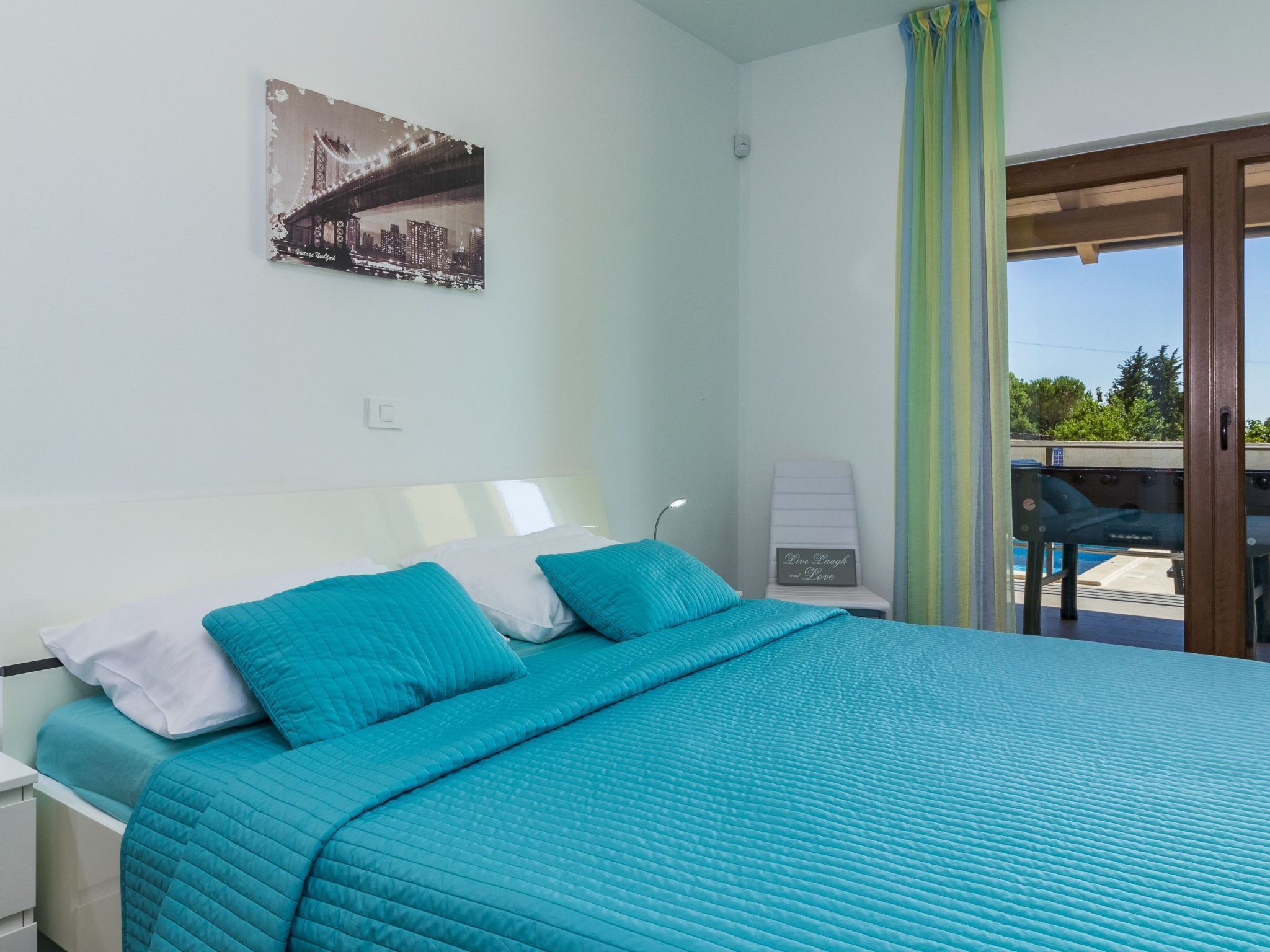 A bedroom with a bed, a nightstand, a wall-mounted lamp, a chair, and a view of a pool and greenery outside.