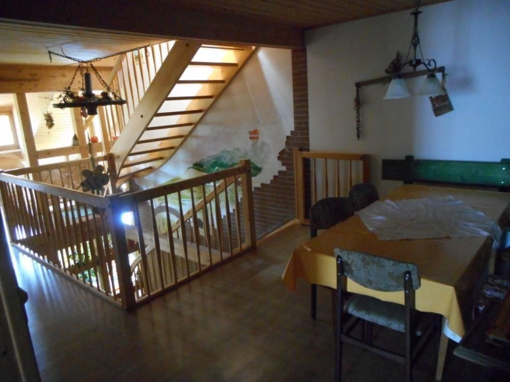 A room with a wooden staircase, a bed with a green headboard, a dining table with chairs, and a hanging light fixture.