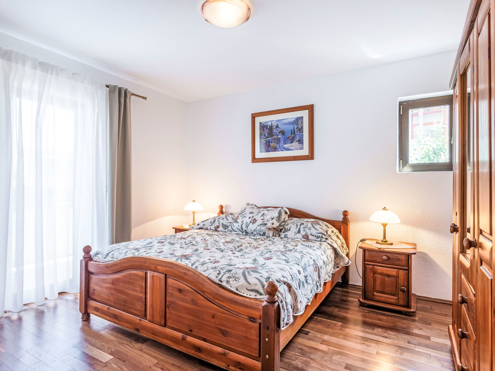 A wooden bed with a floral bedspread, two bedside lamps, a framed picture on the wall, and a wooden wardrobe.