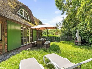 Ein Haus mit einem Reetdach, einer grünen Tür und Fenstern, einem Grill und einem Garten mit Tisch und Stühlen.