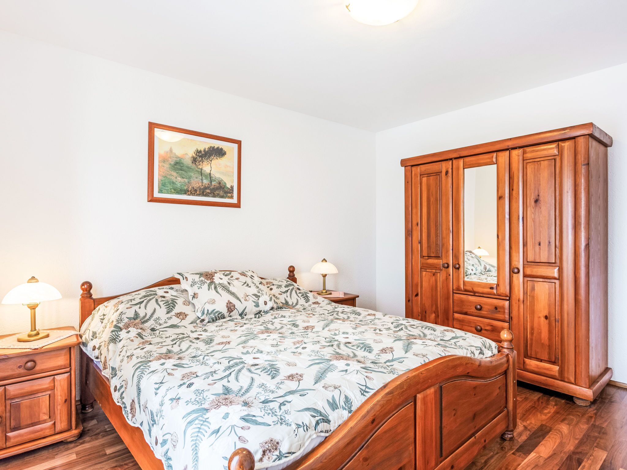 Un dormitorio con cama de madera, mesita de noche con lámpara, armario y un cuadro enmarcado en la pared.