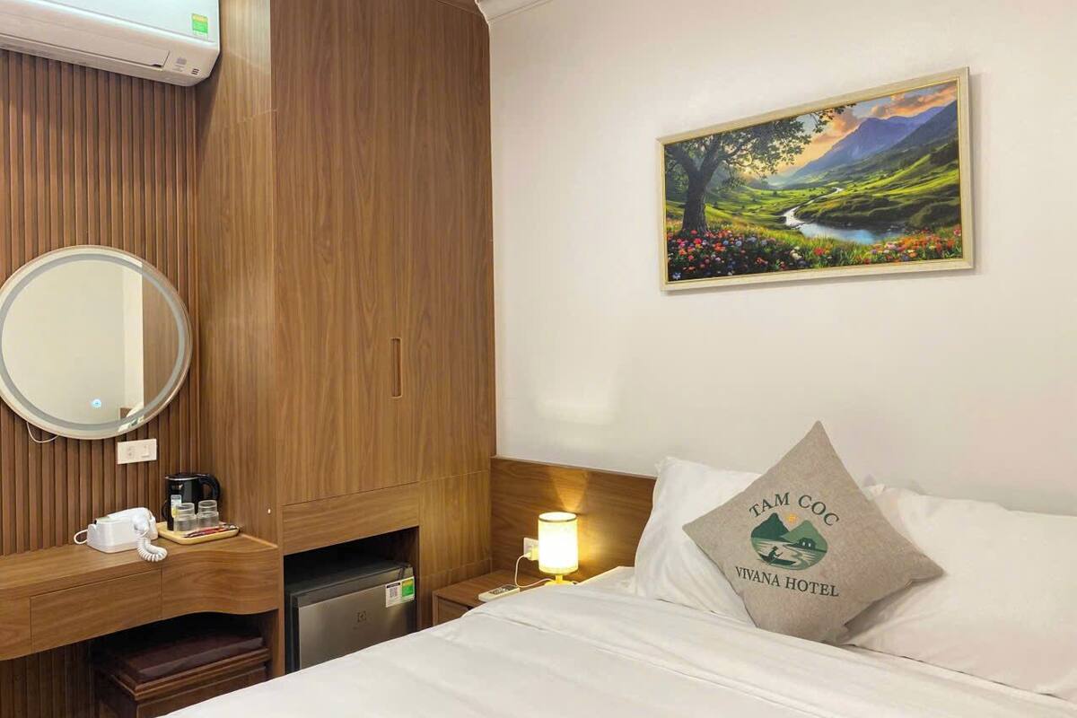 Une chambre d’hôtel avec une armoire en bois, un lit avec un oreiller orné du logo de l’hôtel, une table de chevet avec une lampe et un tableau représentant un paysage accroch é au mur.