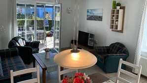 Un saló amb un sofà, una taula amb una làmpada, una televisió, i una vista a través d'una porta de vidre a una zona exterior amb una taula i caires.