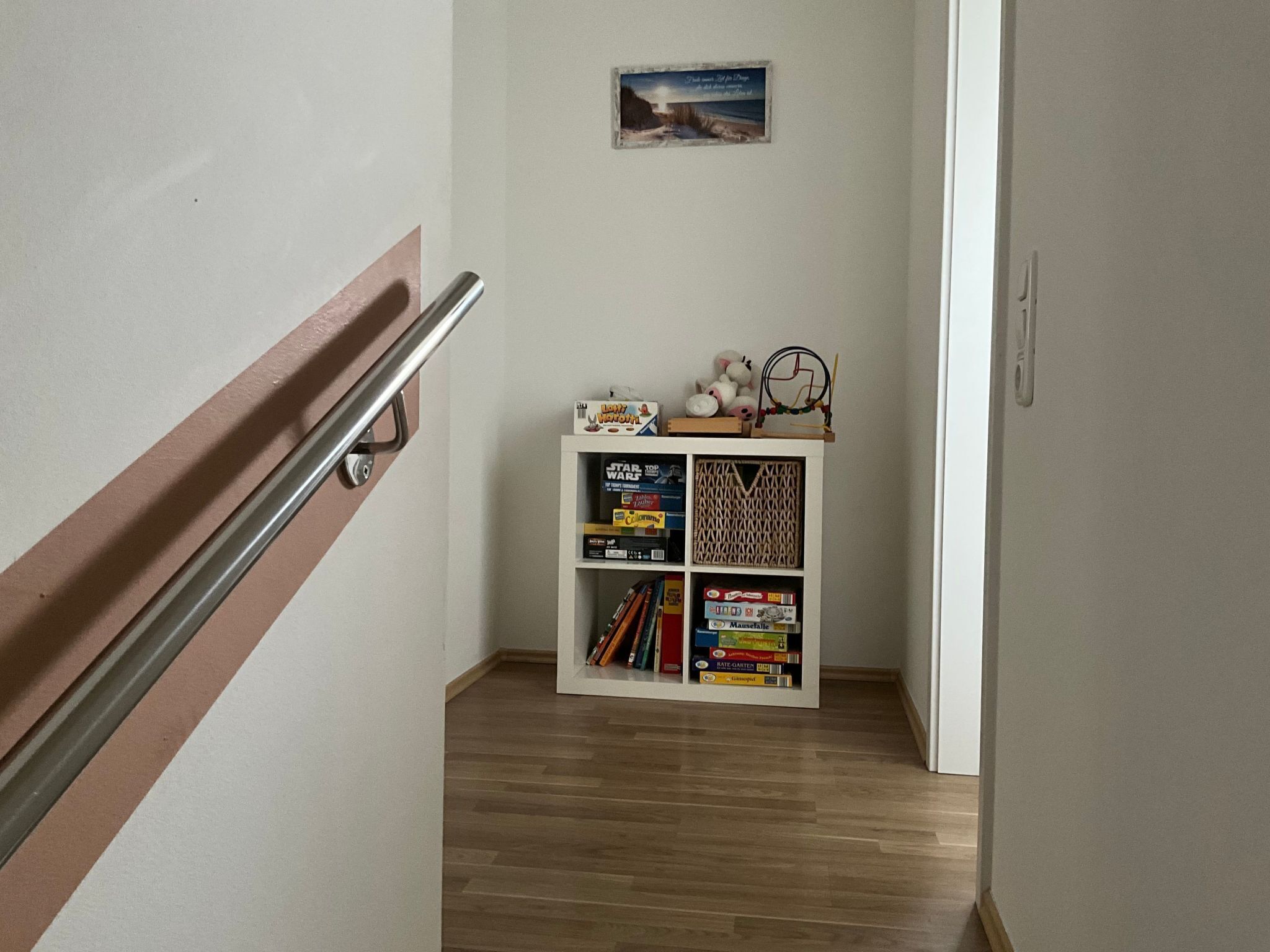 Ein Flur mit Holzboden, einem weißen Regal mit Büchern und Spielzeug sowie einem kleinen Bild an der Wand.