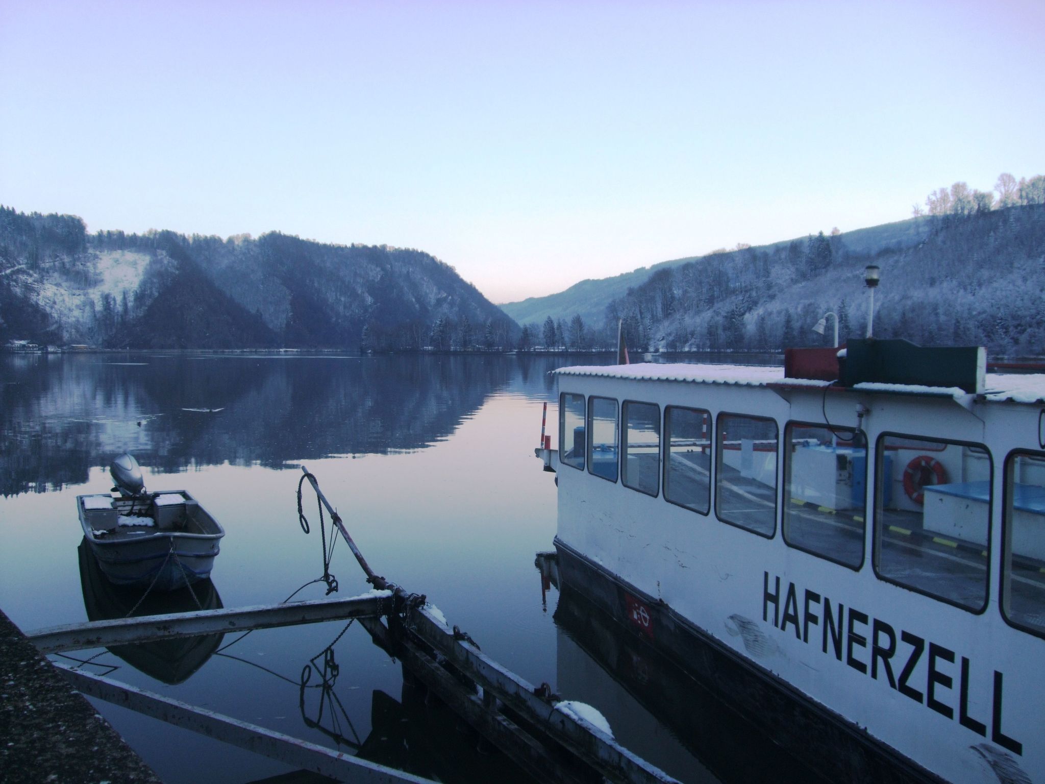Ein Boot liegt an einem ruhigen See, im Hintergrund verschneite Hügel.