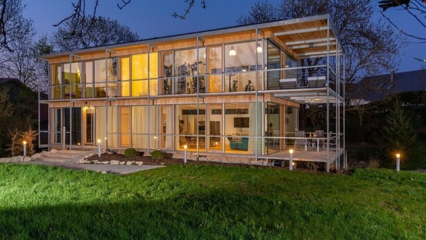 Une maison moderne à deux étages, dotée de grandes fenêtres en verre et d’une terrasse en bois.