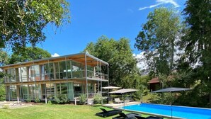 Une maison moderne à deux étages, dotée d’une façade en verre, d’une piscine et d’un espace salon extérieur.