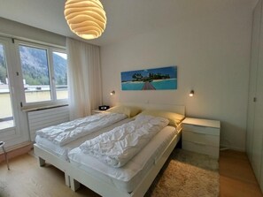 A bedroom with a bed, bedside tables, a painting, and a view of mountains through the window. - Cristal 68 Attika by Interhome (Leukerbad)