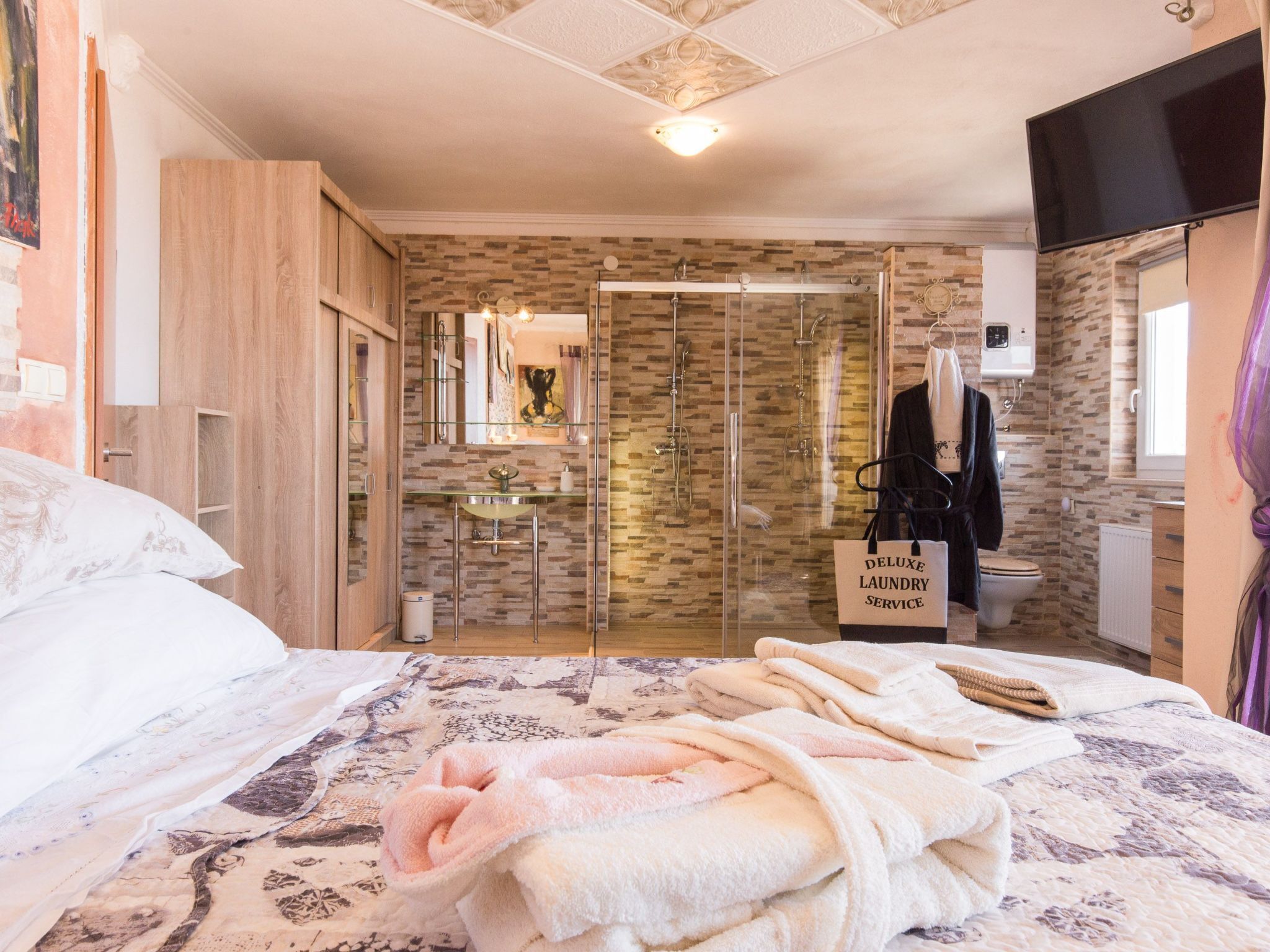 A hotel room with a bed, a bathroom with a shower, a sink, and a mirror. There is a sign for 'Deluxe Laundry Service' in the bathroom.