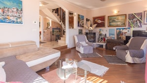 Un salón con un sofá, sillones, una mesa de centro de cristal y una chimenea. Hay cuadros en las paredes y una escalera que lleva a otra habitación.