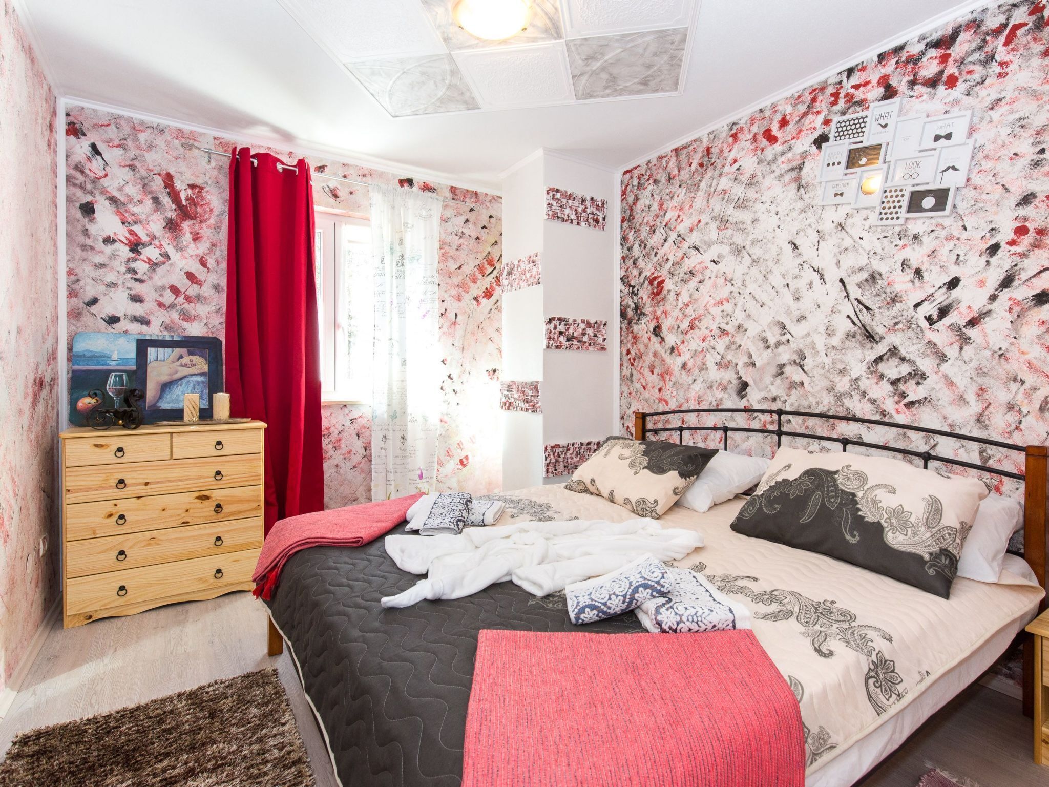 A bedroom with a bed, a wooden dresser, a red curtain, and a wall with a floral pattern.