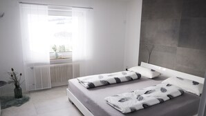 Une chambre moderne avec un grand lit, une fenêtre avec des rideaux légers, un radiateur et une petite plante.