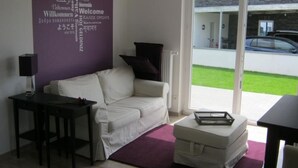 Een moderne woonkamer met een witte bank, een klein tafeltje en een paars vloerkleed. Een woordwolk op de muur toont verschillende welkomstboodschappen in meerdere talen.