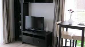 Een moderne woonkamer met een flatscreen televisie op een zwarte tv-meubel, een witte vaas met bloemen op een tafel, en uitzicht op groen buiten.