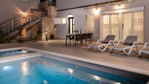Una piscina con sillones de descanso y zona de comedor, rodeada por una casa moderna con fachada de piedra y una escalera.