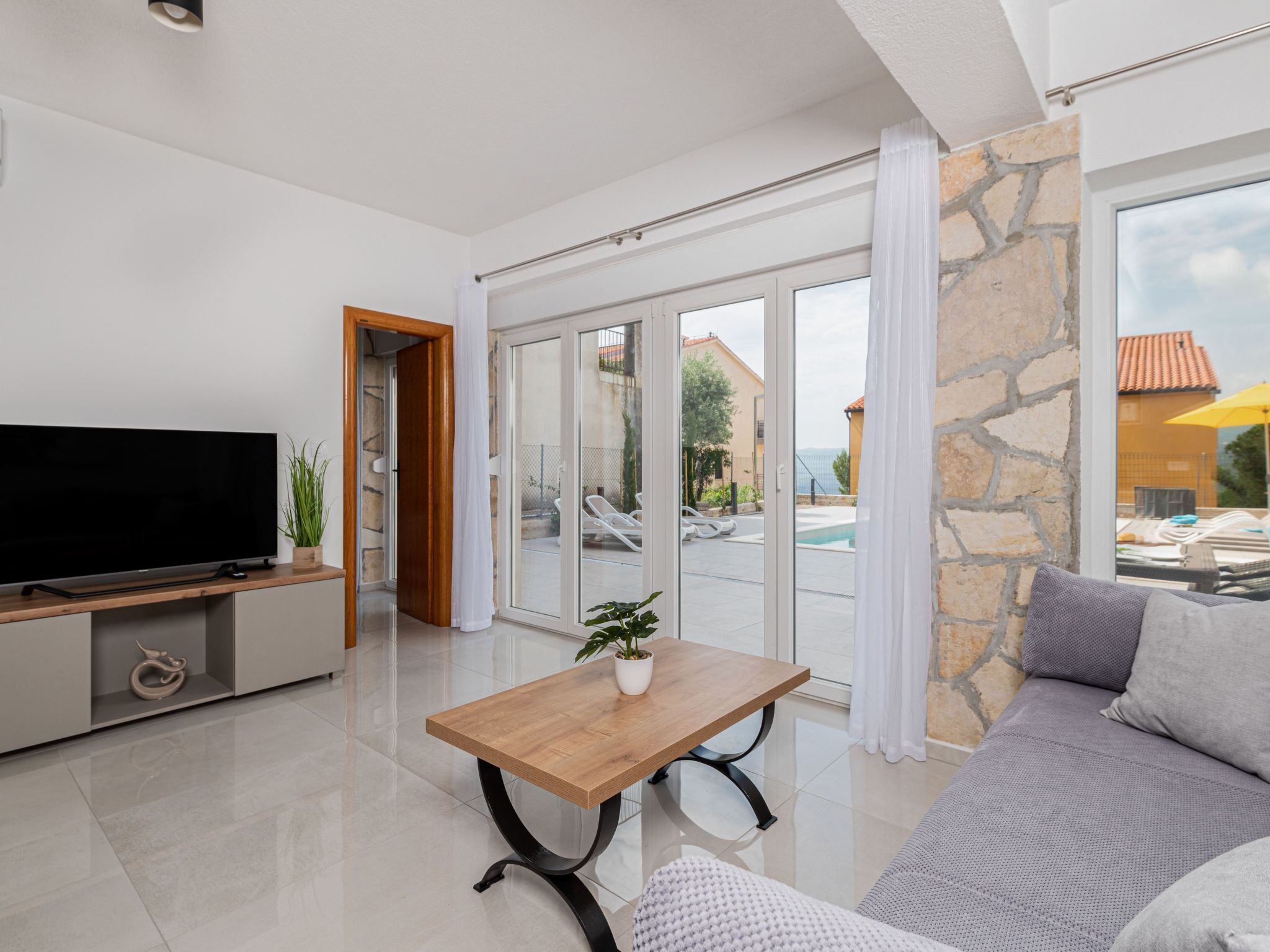 A modern living room with a flat-screen TV, a wooden coffee table with a plant, a grey sofa, and a stone wall.