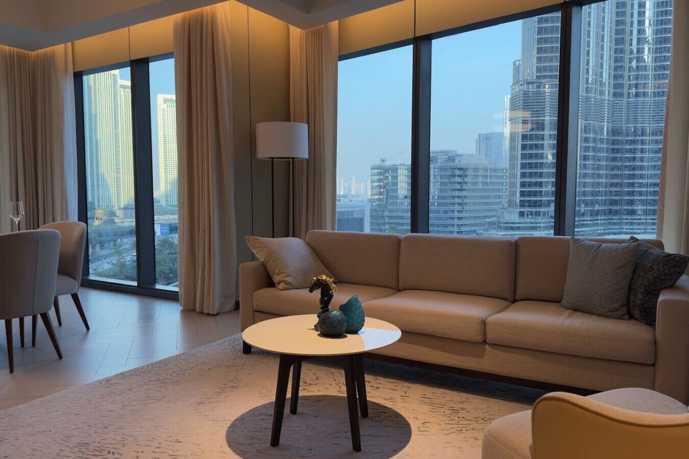 A modern living room with a sofa, armchairs, and a coffee table. Large windows offer a city view.