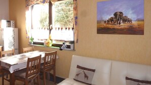 Una habitación con una mesa de comedor, sillas, un sofá, una ventana con cortinas, un cuadro de elefantes y un refrigerador.