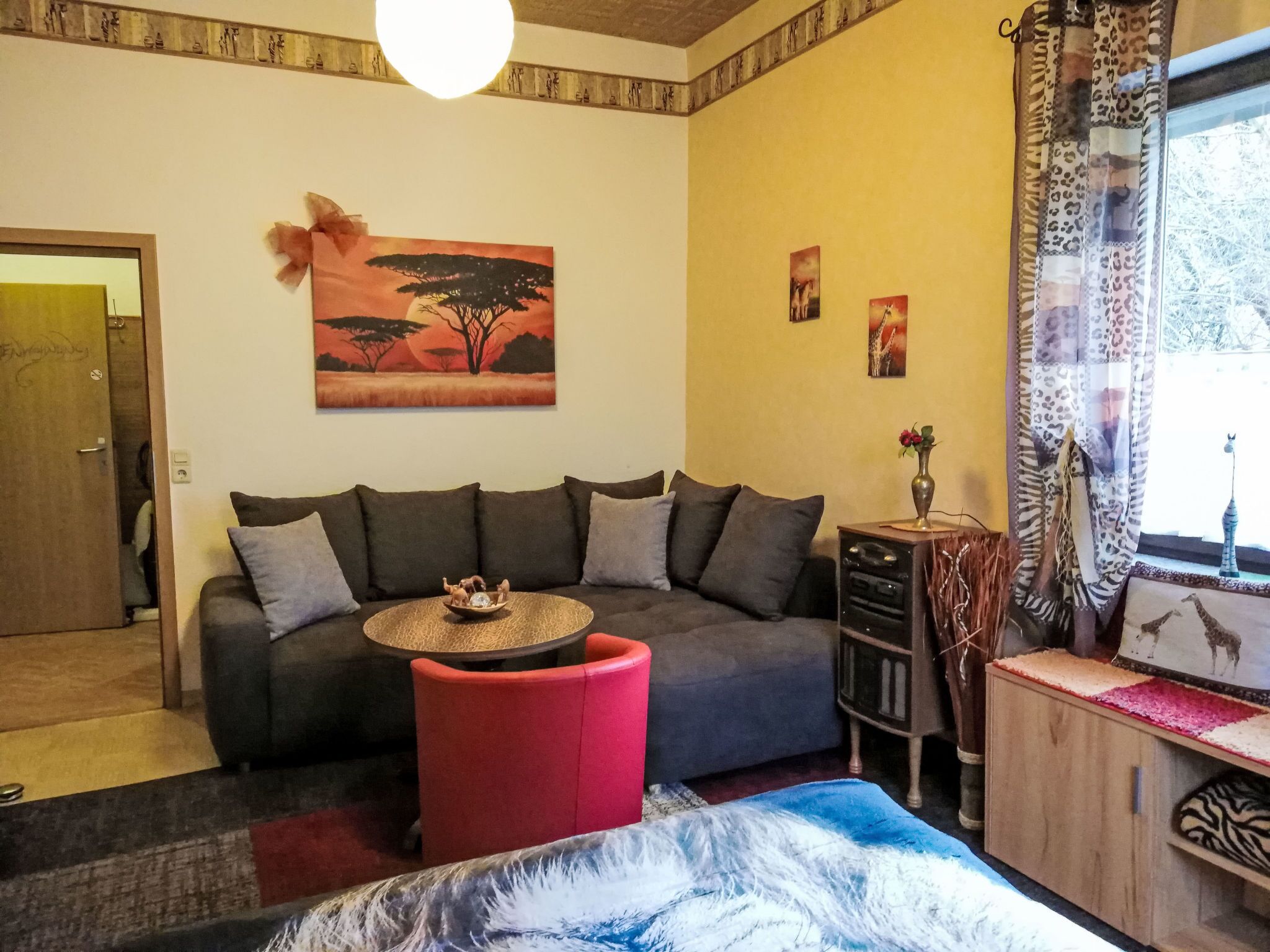 Ein Wohnzimmer mit einer Couch, einem kleinen Tisch, einem roten Hocker, einem Sonnenuntergangsbild und einem Giraffenbild an der Wand.