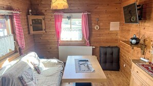 En mysig stuga med en säng, ett bord, en stol, ett fönster med gardiner, ett element och en väggmonterad tv.