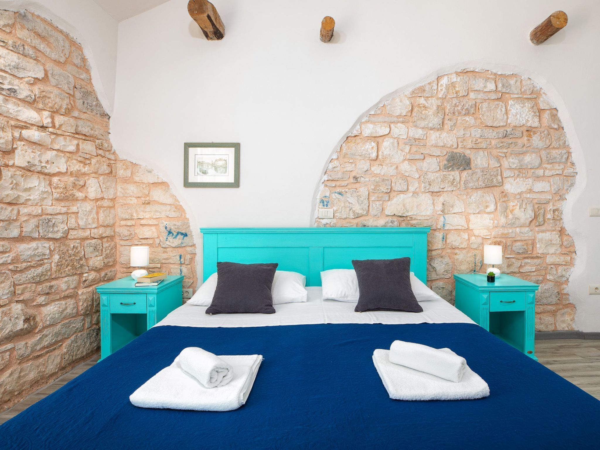 Quarto com parede de pedra, cabeceira turquesa e cama com roupa de cama branca e azul escura.