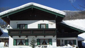 Eine zweistöckige Chalet mit schneebedecktem Dach und grünen Fensterläden, eingebettet in eine verschneite Landschaft.