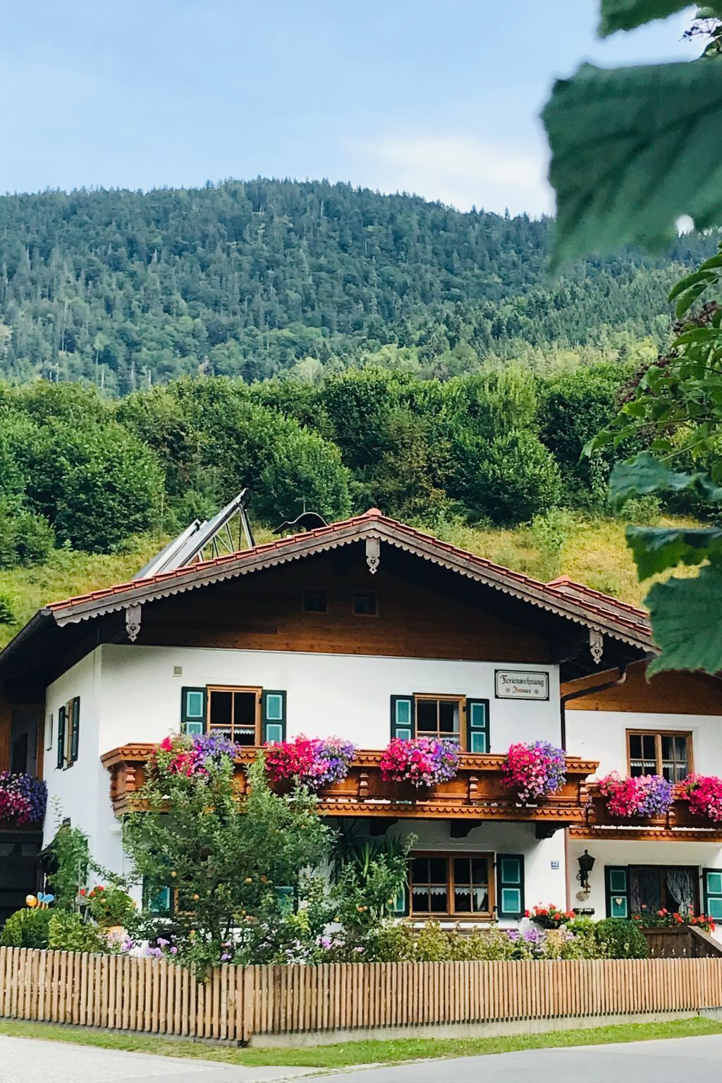 En traditionell chalet med en träbalkong, omgiven av frodig grönska och en skogbeklädd bergssluttning i bakgrunden.