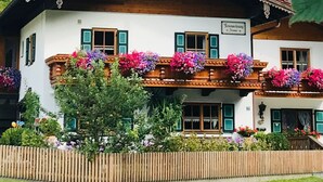 Ein traditionelles Chalet mit Holzbalkon, umgeben von üppiger Vegetation und einer bewaldeten Berglandschaft.
