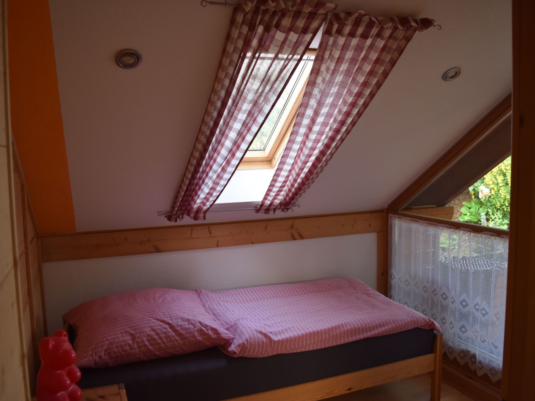 Une petite chambre avec un lit simple, un plafond en pente, un puits de lumière et une fenêtre ornée de rideaux en dentelle.