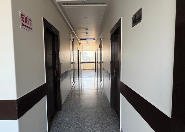 A long, narrow hallway with doors on both sides, a sign indicating an exit, and a 'No Smoking' sign. - Hotel Girraj Inn (Govardhan)