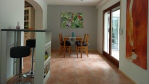 Uma área de refeições com mesa e cadeiras, uma cozinha com bancada e bancos, e uma janela grande com vista.