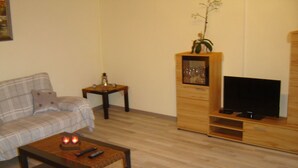 A living room with a sofa, a coffee table with remotes, a TV on a wooden stand, and a small table with a vase and candle. - Santa Maria by Interhome (Nettersheim)