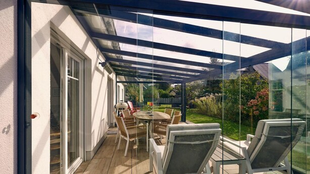 A covered patio with a glass enclosure, a wooden table, and white chairs. - Schönes Ferienhaus in Brochterbeck mit Garten by Interhome (Tecklenburg)