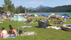 Ein belebter Seeuferbereich mit Menschen, die im Gras entspannen, bunten Sonnenschirmen und einem Berg im Hintergrund.