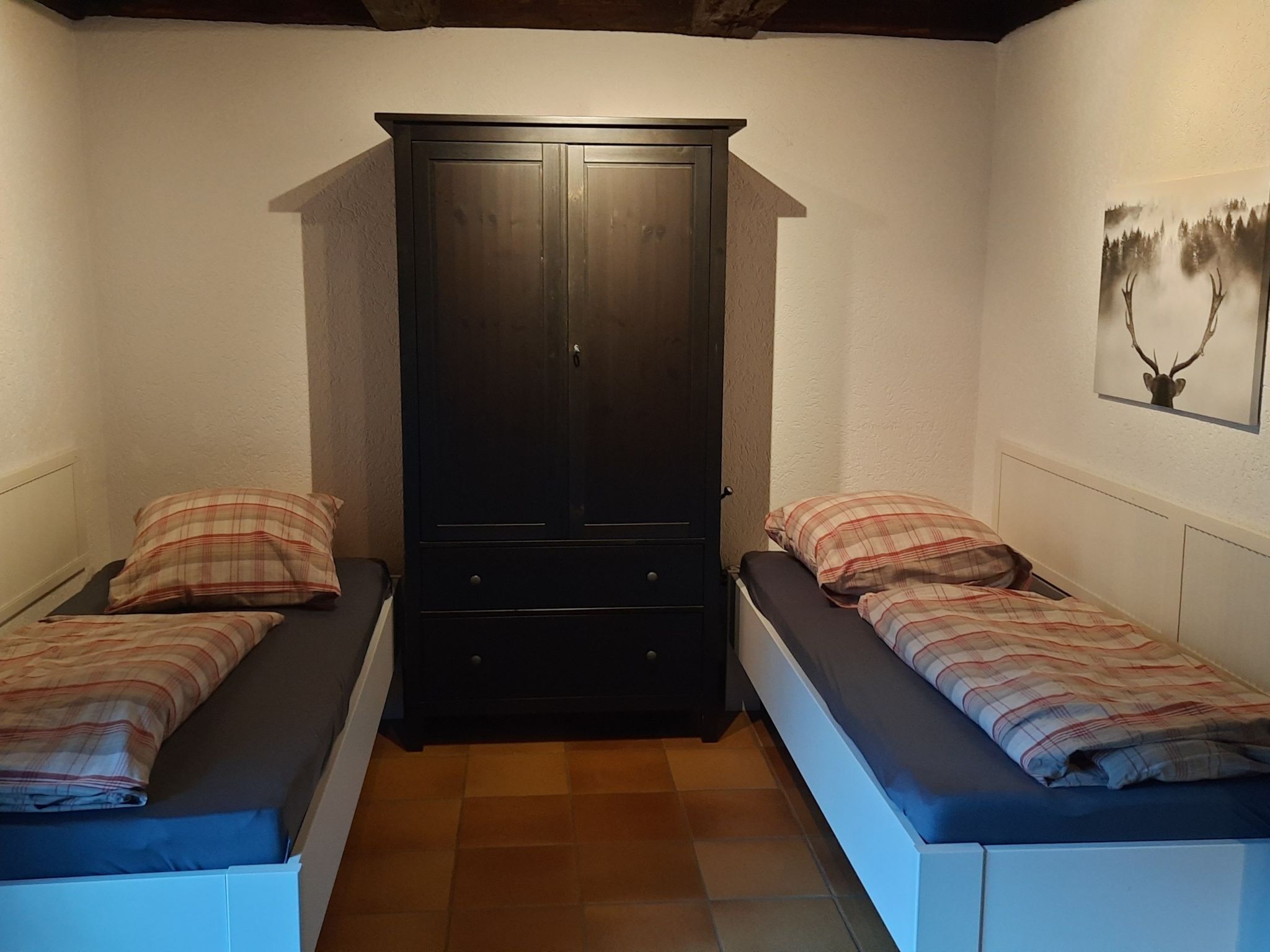 Une petite pièce avec deux lits simples, une armoire en bois sombre et un tableau représentant une tête de cerf, encadré, accroché au mur.