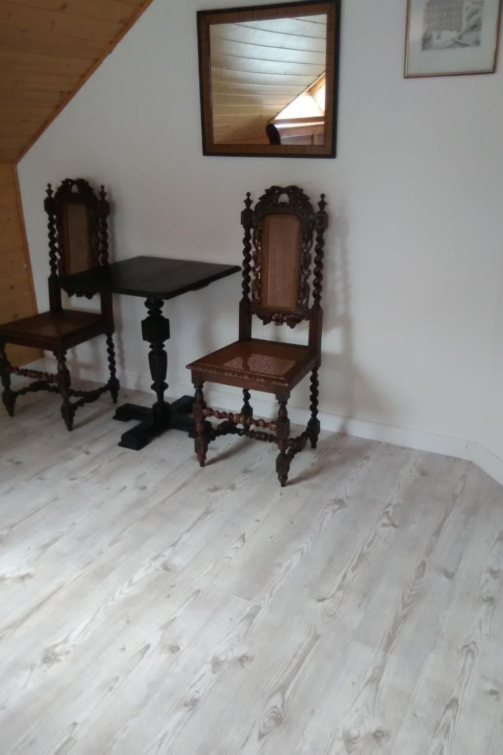 A room with a wooden floor, a dark wooden table, two ornate chairs, and a framed mirror on the wall.