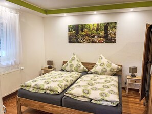 Una cama doble con ropa de cama floral, una mesita de noche con lámpara, un cuadro de un bosque y suelo de madera.