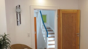 Ein Zimmer mit Holzboden, einem Korbstuhl, einem Glastisch mit Dekoration und einer Holztür sowie einer Treppe mit weißen Geländern.