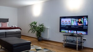 Uma sala de estar moderna com uma TV de tela plana na parede, um sofá cinza, um banquinho preto, uma planta em vaso e uma mesa pequena com tampo de vidro.
