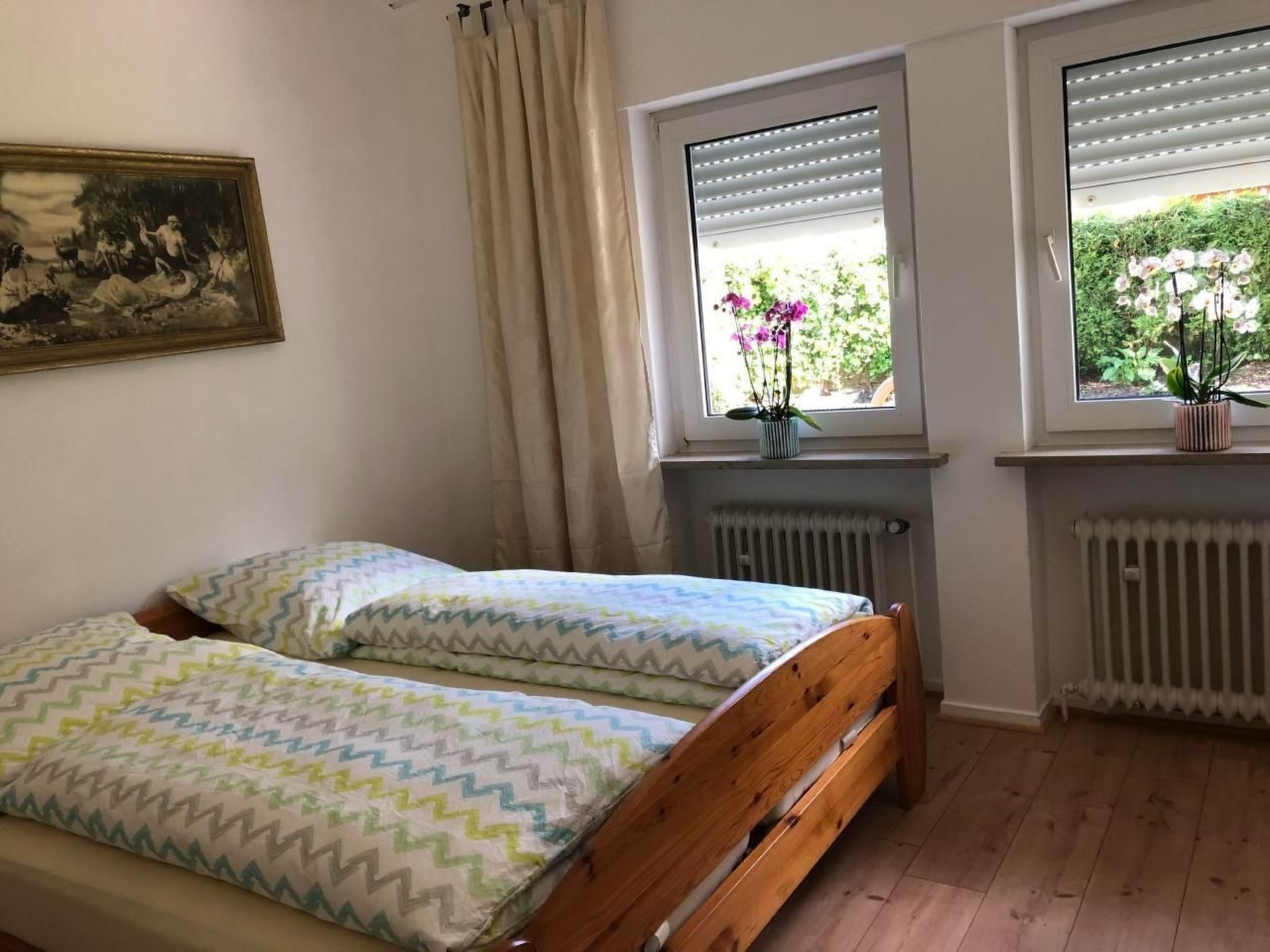 Une chambre à coucher avec un lit en bois, deux fenêtres équipées de stores, un radiateur et un tableau accroché au mur.