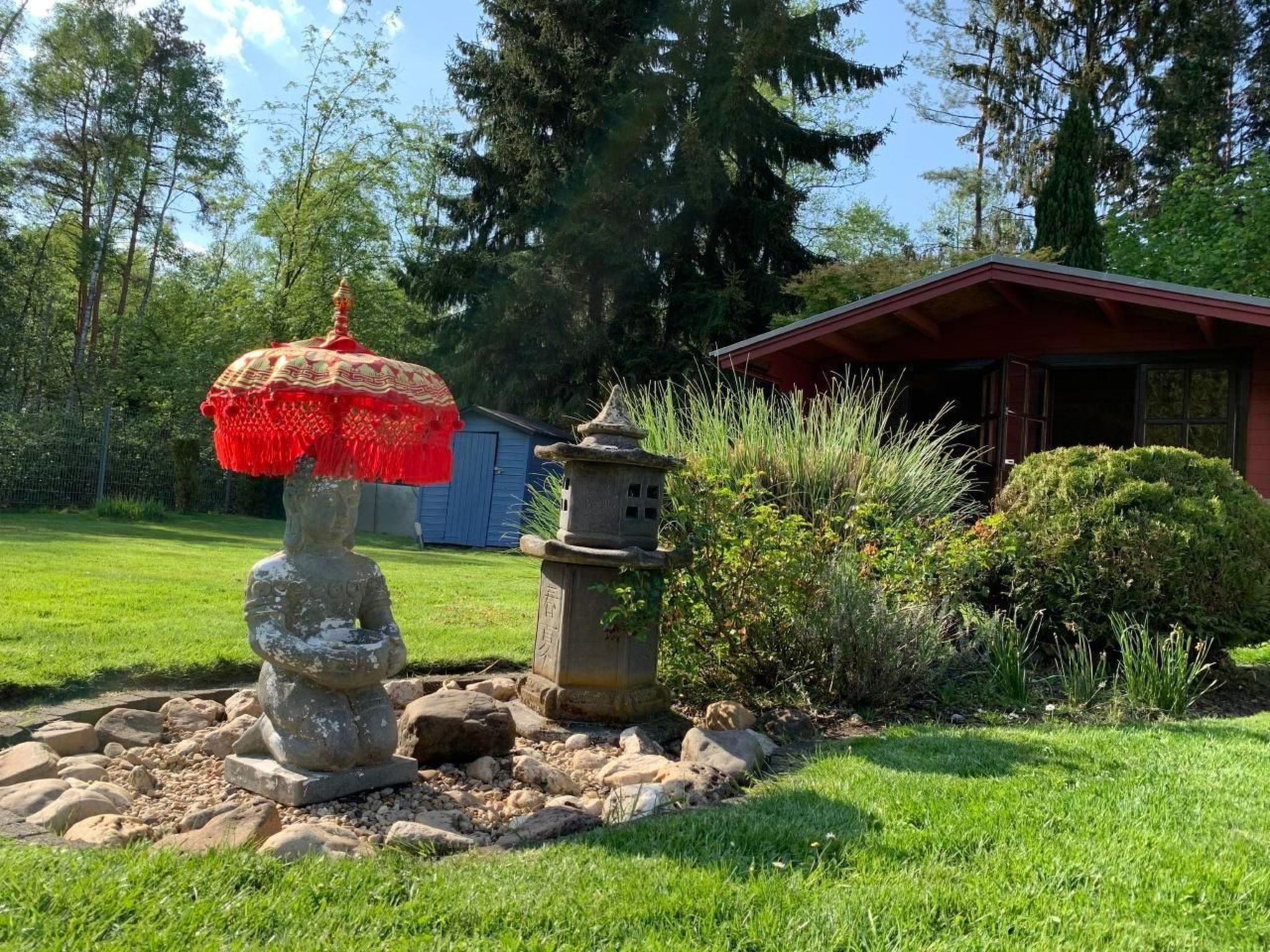 A stone statue with a red umbrella, a stone lantern, and a small shrine in a garden with a red building in the background.