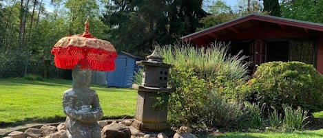 A stone statue with a red umbrella, a stone lantern, and a small shrine in a garden with a red building in the background.