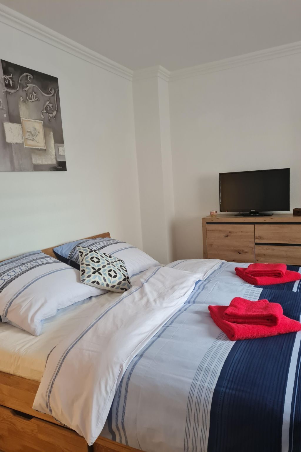 Ein ordentlich bezogenes Bett mit gestreifter Bettwäsche, ein Fernseher auf einem Holzboden und ein Wandbild über dem Bett.