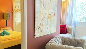 Een kamer met een bed, een gestoffeerde fauteuil, een schilderij aan de muur en een raam met gordijnen.