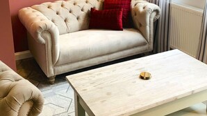 Een woonkamer met een beige bank, twee abstracte schilderijen, een witte salontafel en een raam met gordijnen.