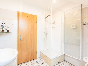 Een badkamer met een houten deur, een wit wastafelmeubel, een douchecabine van glas en een betegelde vloer.