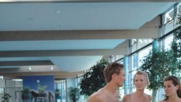 Ein Schwimmbecken mit einer Metallleiter und einer Person in blauen Badehosen am Beckenrand.