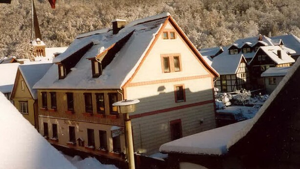 A traditional chalet-style building with a steeply pitched roof, surrounded by snow-covered houses and a forested hill in the background. - Gästehaus Tröbs by Interhome (Ilfeld)