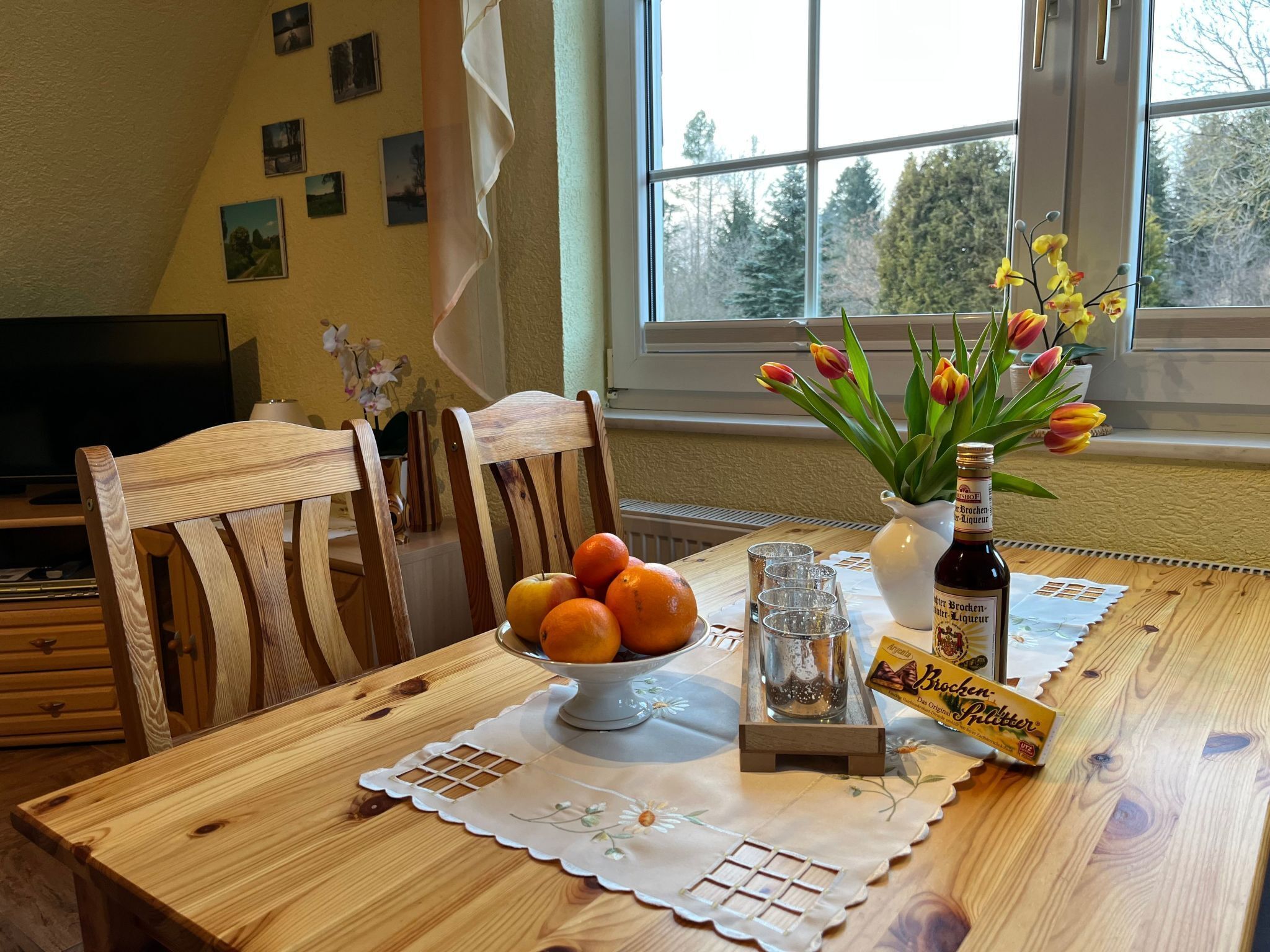 A wooden dining table with a bowl of oranges, a bottle of Bräu-Bier, and tulips.