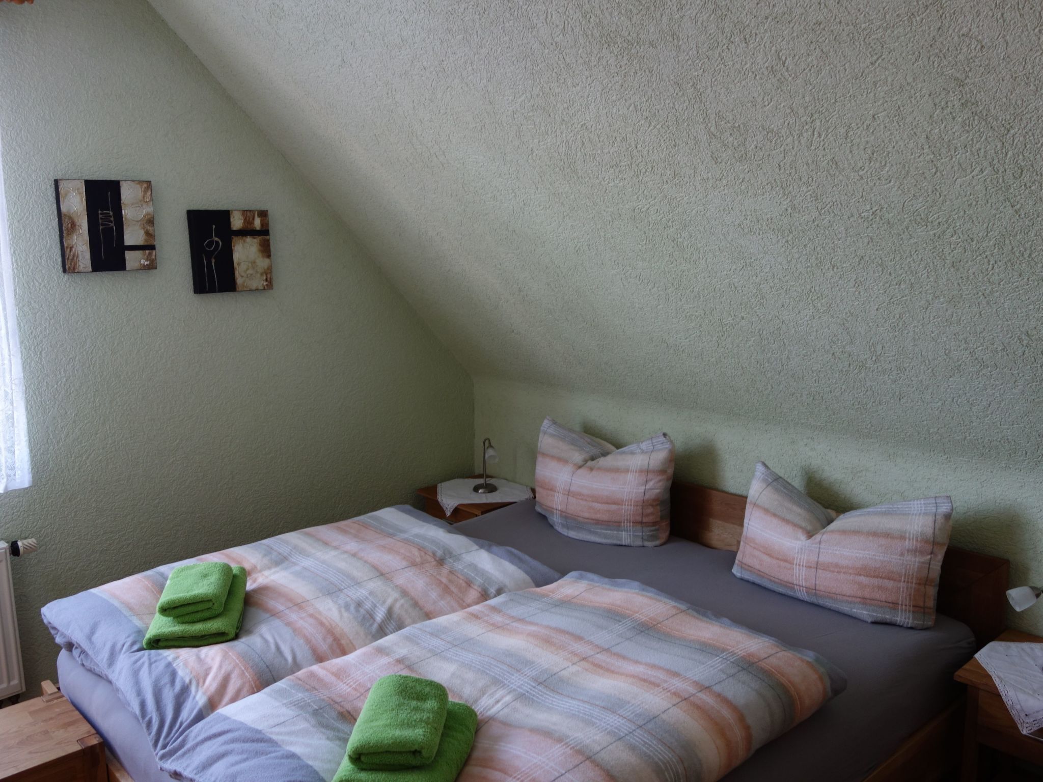 A bedroom with two beds, each with a plaid bedspread and green towels. There are two wall art pieces and a nightstand with a lamp.